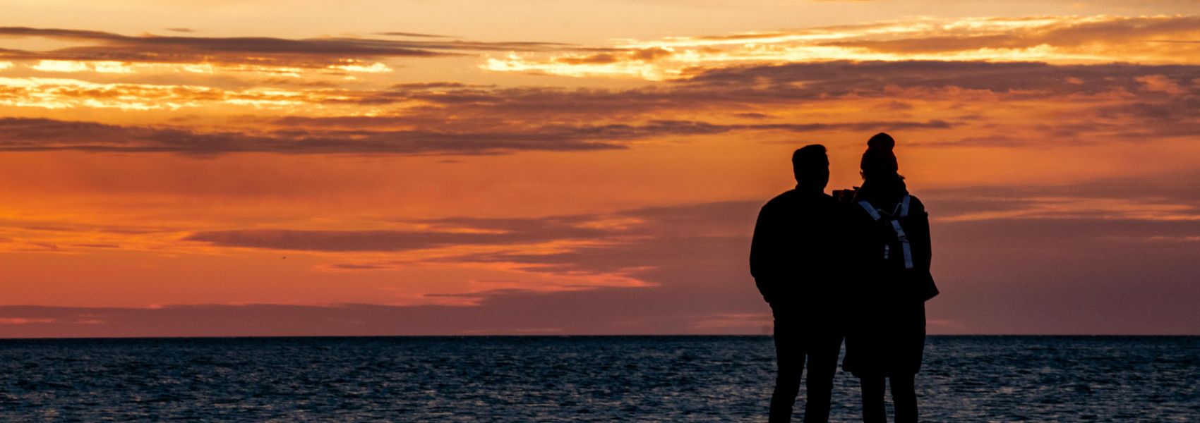 Dark sillouhette of a couple from behind, standing on a beach and staring out over the sea during a deep orange sunset
