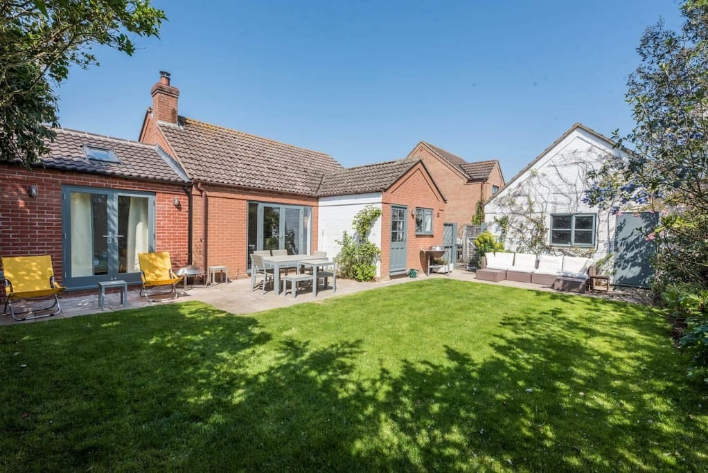 A large red brick home in the background with a bright green grass garden in the foreground complete with garden furniture