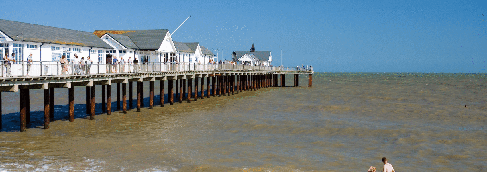 Long wooden pier to the left over the ocean, with sand and people playing in the foreground and blue sky in the background