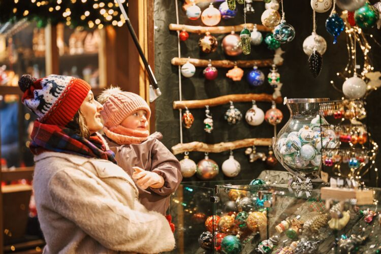 A mother holding her baby, both in winter cothing, looking happily at a wooden stall selling colourful Christmas decorations