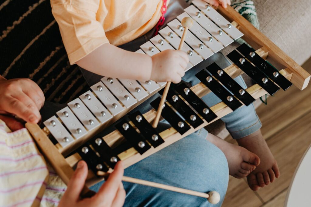 Close up of a small zylophone on the lap of a toddler who is playing it while an adult's hands help