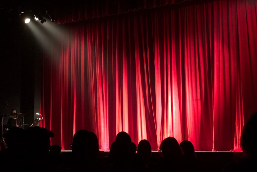 Red curtains across a theatre stage with a bright spotlight on their centre, and the shadows of the backs of people's heads in the audience