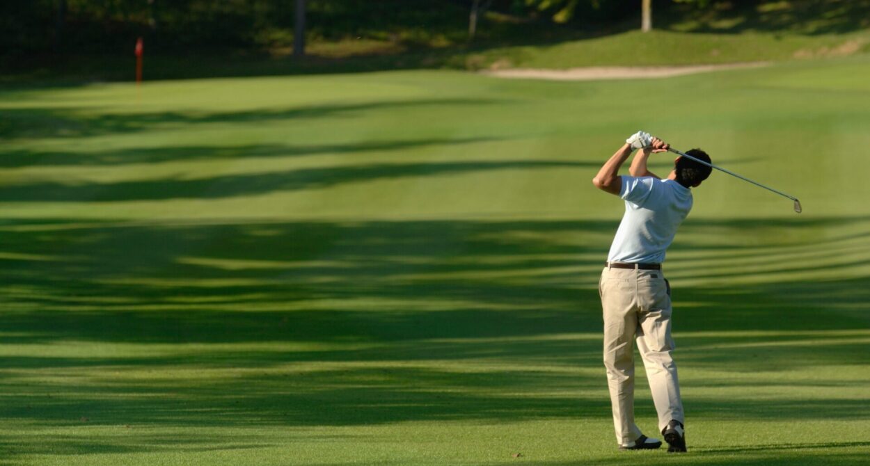 Man mid-swing playing golf on a lush green golf course with stretching tree shadows