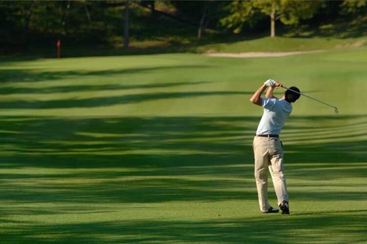 Man mid-swing playing golf on a lush green golf course with stretching tree shadows