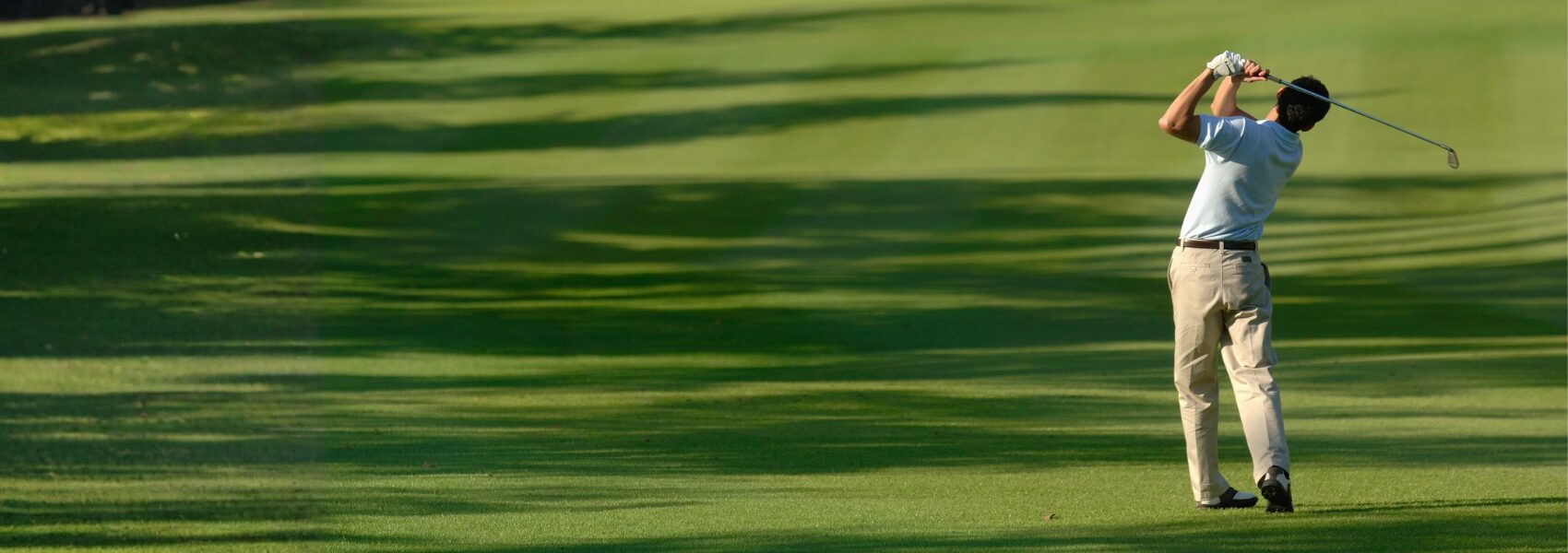 Man mid-swing playing golf on a lush green golf course with stretching tree shadows