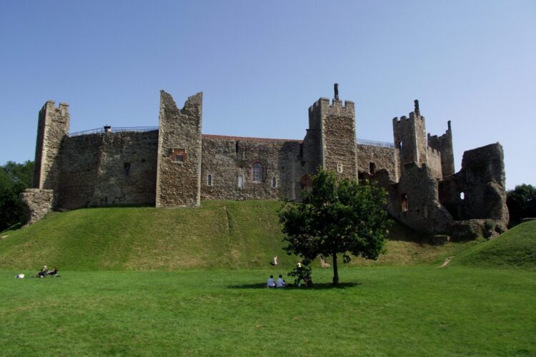 The side of a large stone brick castle on top of a sloping green grassy hill
